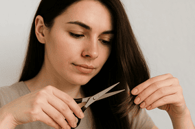 En esta imagen vemos a una mujer joven cortando cuidadosamente las puntas de su cabello con unas tijeras pequeñas. Ella se muestra concentrada, lo que transmite la importancia del cuidado personal. Esta escena es perfecta para ilustrar la pregunta ¿Cuántas veces debo cortar el cabello para mantenerlo sano?, ya que representa un momento de mantenimiento capilar que ayuda a evitar las puntas abiertas y a conservar el cabello fuerte y saludable. La iluminación suave y el fondo neutro resaltan el enfoque en la acción de cortar el cabello, haciendo énfasis en la rutina esencial para quienes buscan un cabello bien cuidado.
