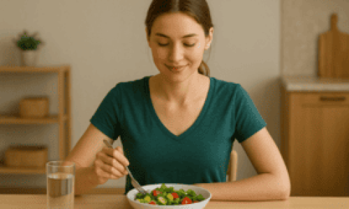 Una mujer joven con expresión tranquila y rostro saludable está sentada en una mesa de madera clara, disfrutando de una ensalada fresca. Lleva una camiseta verde y sostiene un tenedor mientras mira su plato con satisfacción. A su lado hay un vaso de agua, y el fondo muestra una cocina moderna con decoración simple. Esta imagen representa perfectamente el concepto de una alimentación equilibrada y responde visualmente a la pregunta ¿Qué Dieta Debo Seguir para Perder Peso con Salud?, transmitiendo bienestar, simplicidad y hábitos saludables.

