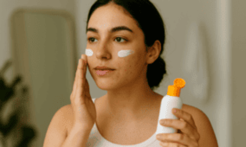 Una mujer joven con piel grasa y algunas imperfecciones visibles se aplica protector solar en el rostro frente a un espejo. Lleva una camiseta sin mangas blanca y sostiene un envase de protector solar con tapa naranja brillante. Tiene una expresión tranquila y concentrada mientras distribuye el producto en sus mejillas. La imagen transmite un momento de autocuidado y es ideal para ilustrar el tema ¿Qué protector solar es mejor para piel grasa?, destacando la importancia de elegir productos adecuados para este tipo de piel.

