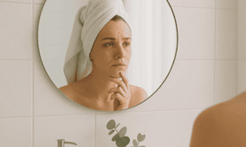 Una mujer joven con la piel clara está frente al espejo redondo de un baño minimalista, envuelta en una toalla blanca sobre la cabeza. Su expresión es pensativa, como si se preguntara ¿cómo cuidar la piel del rostro?. Frente al lavamanos se ven detalles delicados como un dispensador de jabón beige, un pequeño frasco de vidrio con ramas de eucalipto y frascos cosméticos. La iluminación natural y suave del espacio transmite calma y cuidado personal. Esta imagen representa ese momento íntimo en que una persona evalúa su piel con atención y busca mejorar su rutina facial.









